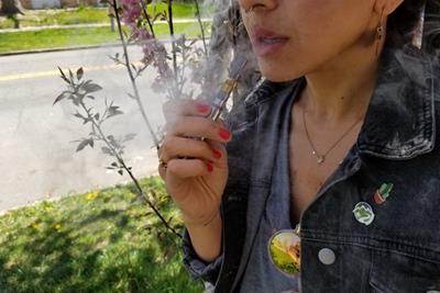 Woman smoking a vape pen in front of tree, with denim jacket on.